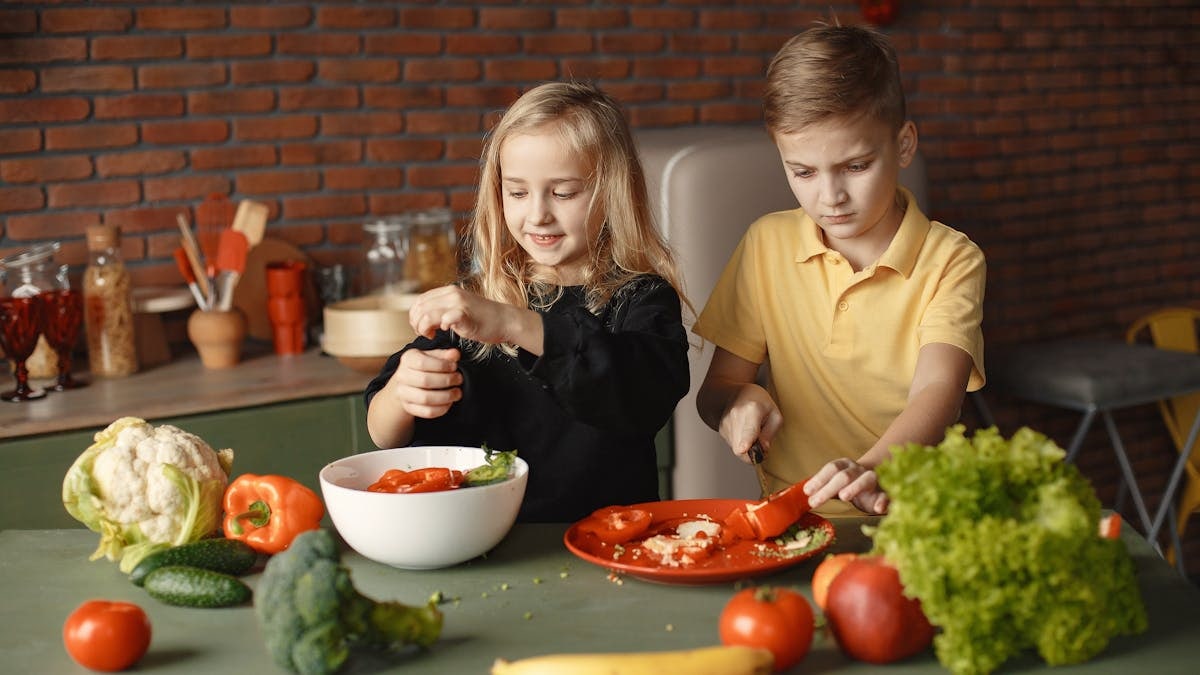 Serious boy cutting pepper for vegetable salad while standing near happy sister seasoning ingredients put into deep bowl in loft style kitchen