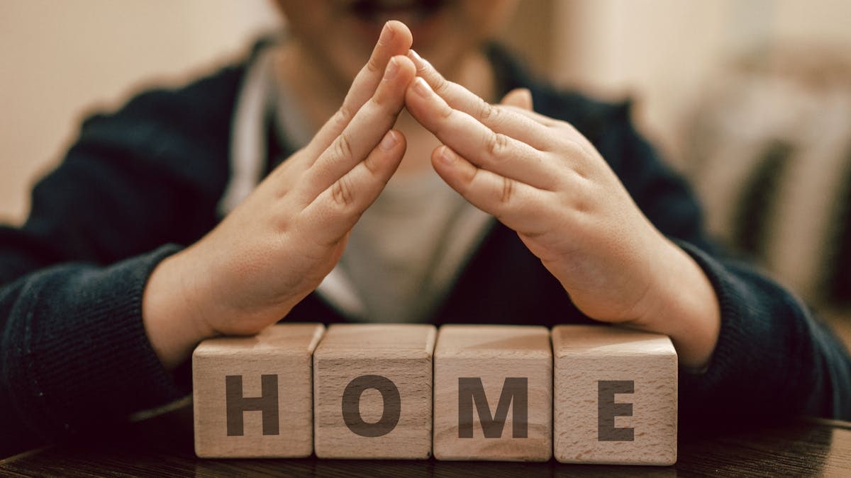 Crop excited kid with mouth opened making roof with hands above wooden cubes with word Home waiting for foster family