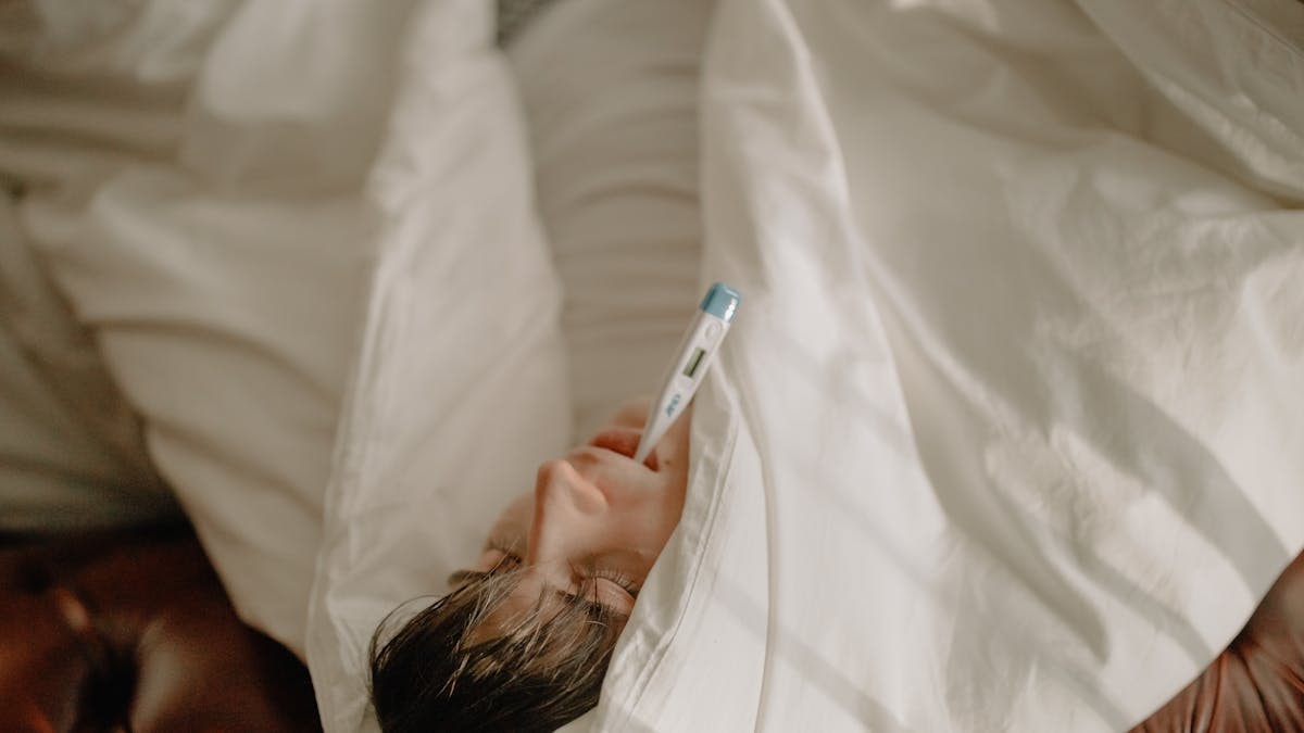 A man lying in bed with a thermometer in his mouth, wrapped in cozy bedding, indicating illness or fever.